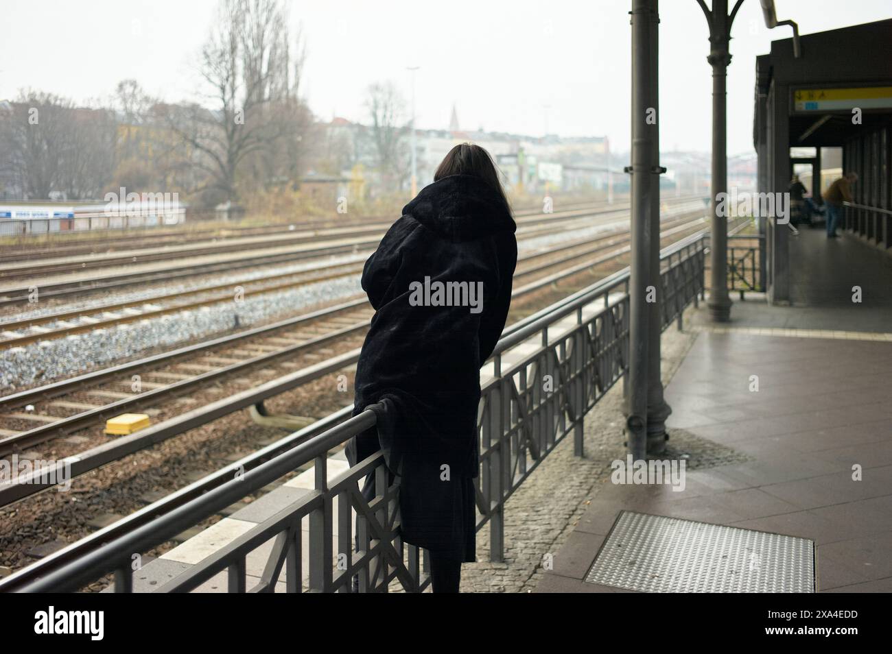 Una persona si trova appoggiata su una ringhiera presso la piattaforma della stazione ferroviaria, osservando i binari in una giornata nuvolosa. Foto Stock