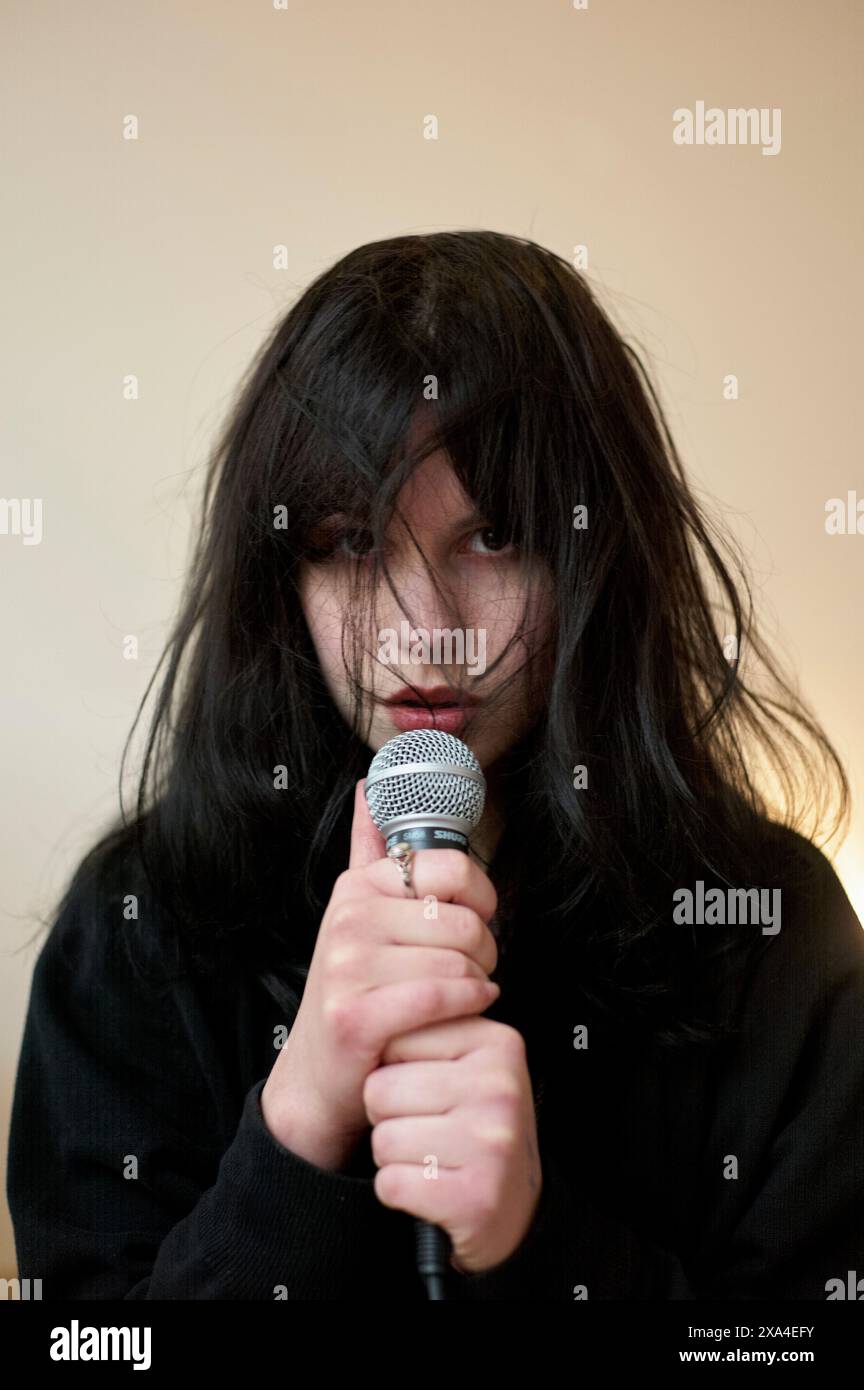 Una persona con i capelli neri disordinati copre parte del viso, tenendo un microfono vicino alle labbra, di fronte a uno sfondo beige semplice. Foto Stock