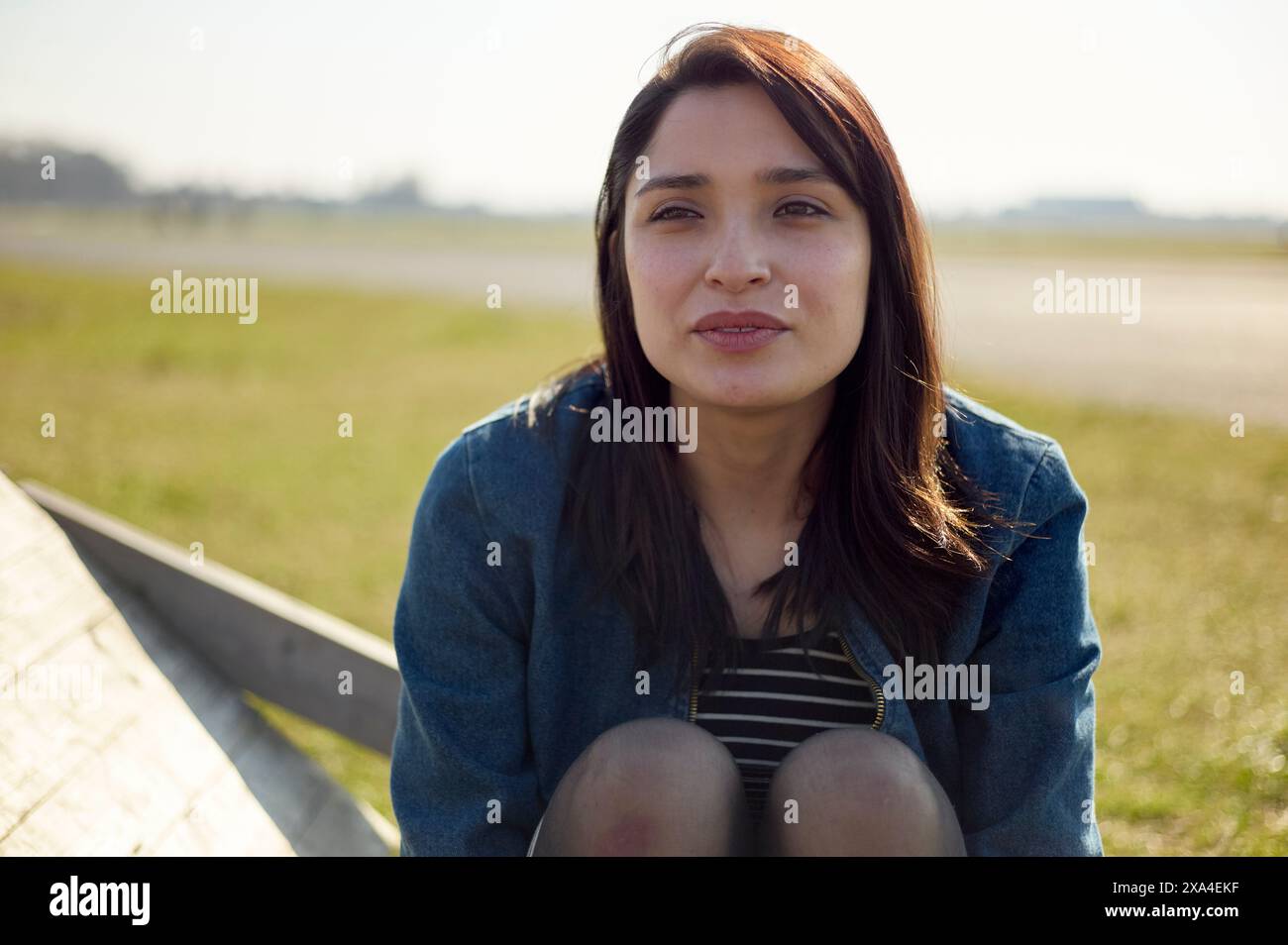 Una donna è seduta all'aperto, strisciando leggermente alla luce del sole, con un sorriso morbido e una postura rilassata. Foto Stock