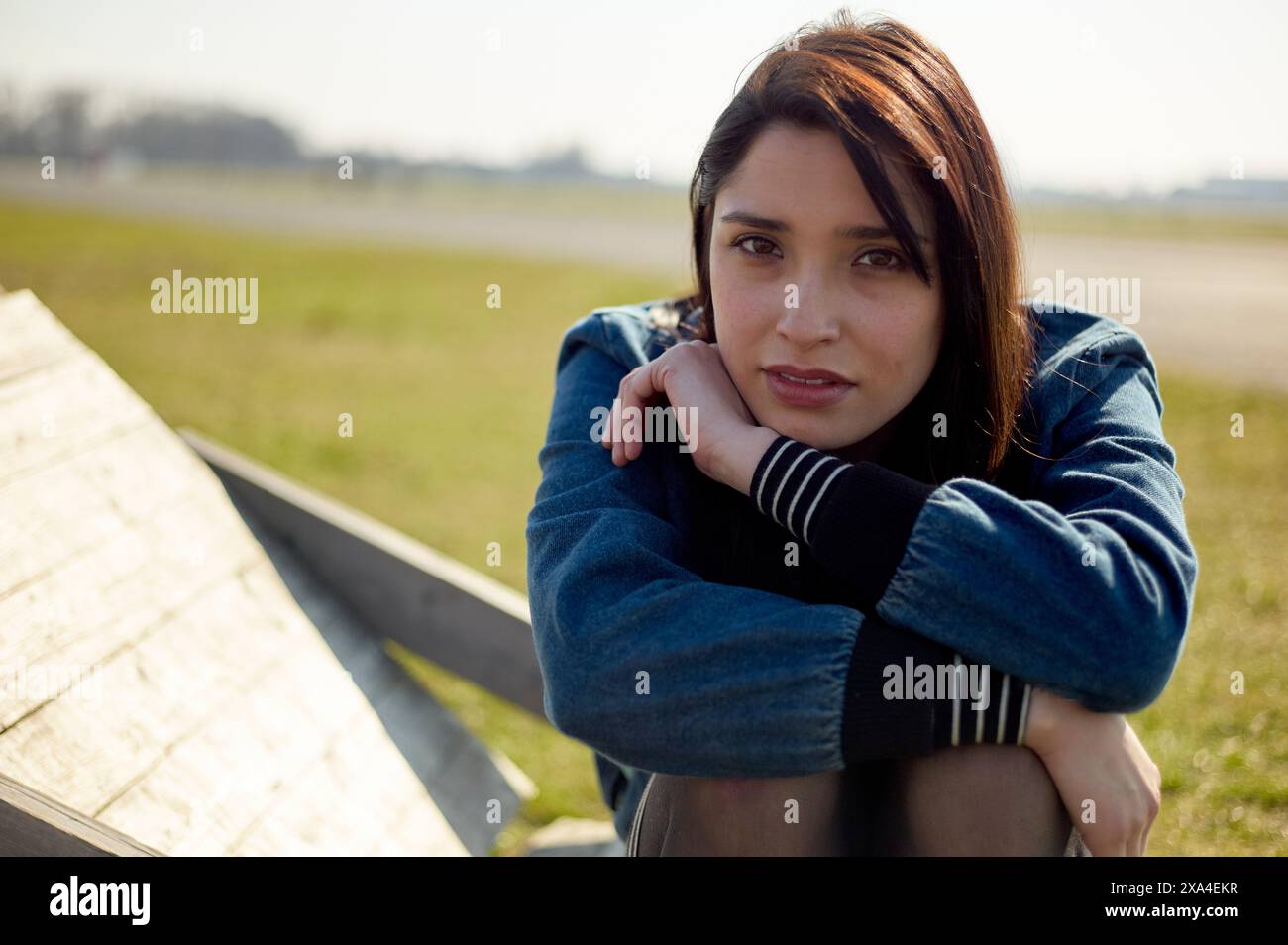 Una donna con capelli castani di media lunghezza siede all'aperto, poggia il mento sulle braccia incrociate, con uno sfondo morbido con erba e un cielo soleggiato. Foto Stock