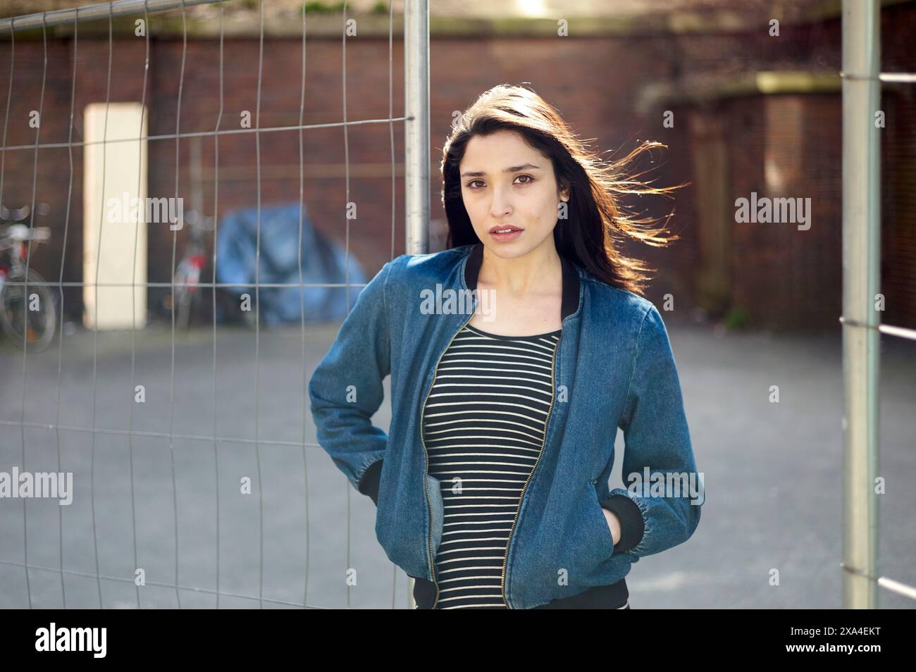 Una giovane donna si trova di fronte a una rete da calcio, i suoi capelli scuri soffiano nel vento, ed è rivestita con una giacca blu sopra una maglia a righe. Foto Stock