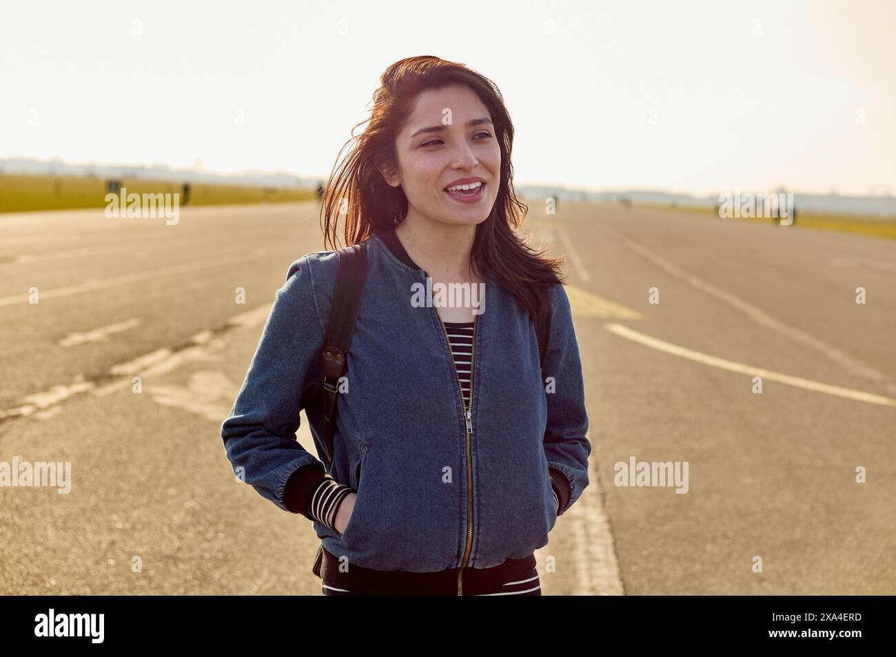 Una donna sorridente si trova su una strada aperta, retroilluminata da una luce solare dorata e soffusa con un cielo limpido alle sue spalle. Foto Stock