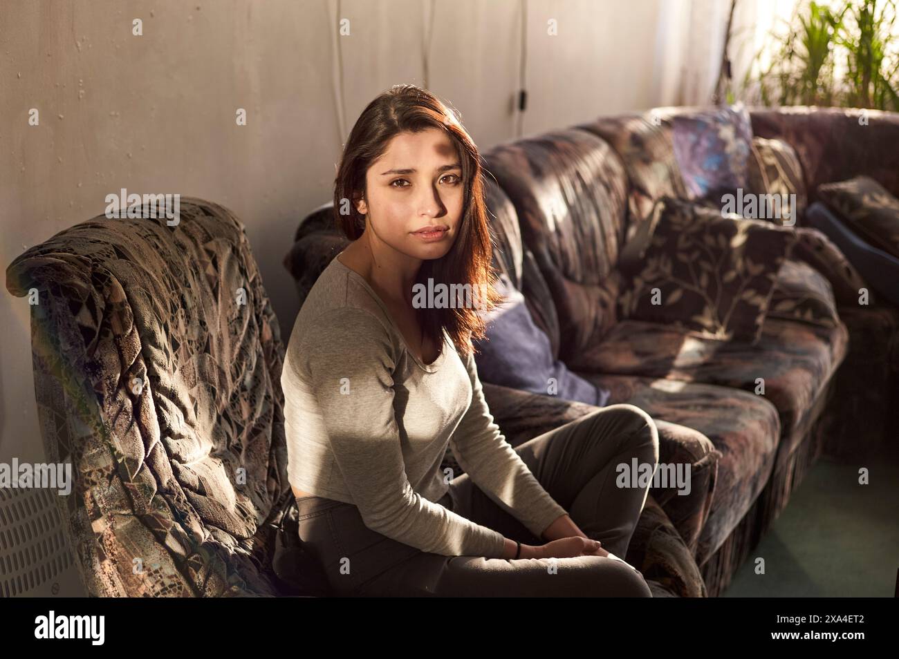 Una giovane donna è seduta su un divano floreale, inondata di luce naturale, con un'espressione penosa sul viso. Foto Stock