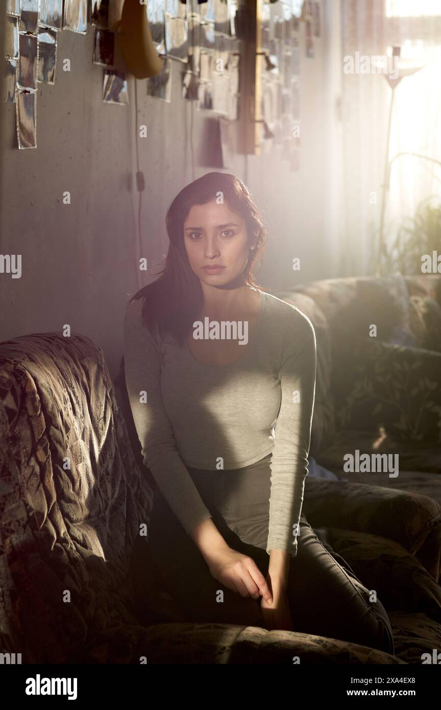 Una donna è seduta su un divano in una stanza illuminata dalla luce naturale, con la luce che crea un effetto caldo e appannato. Appare contemplativa mentre guarda verso la telecamera. Foto Stock