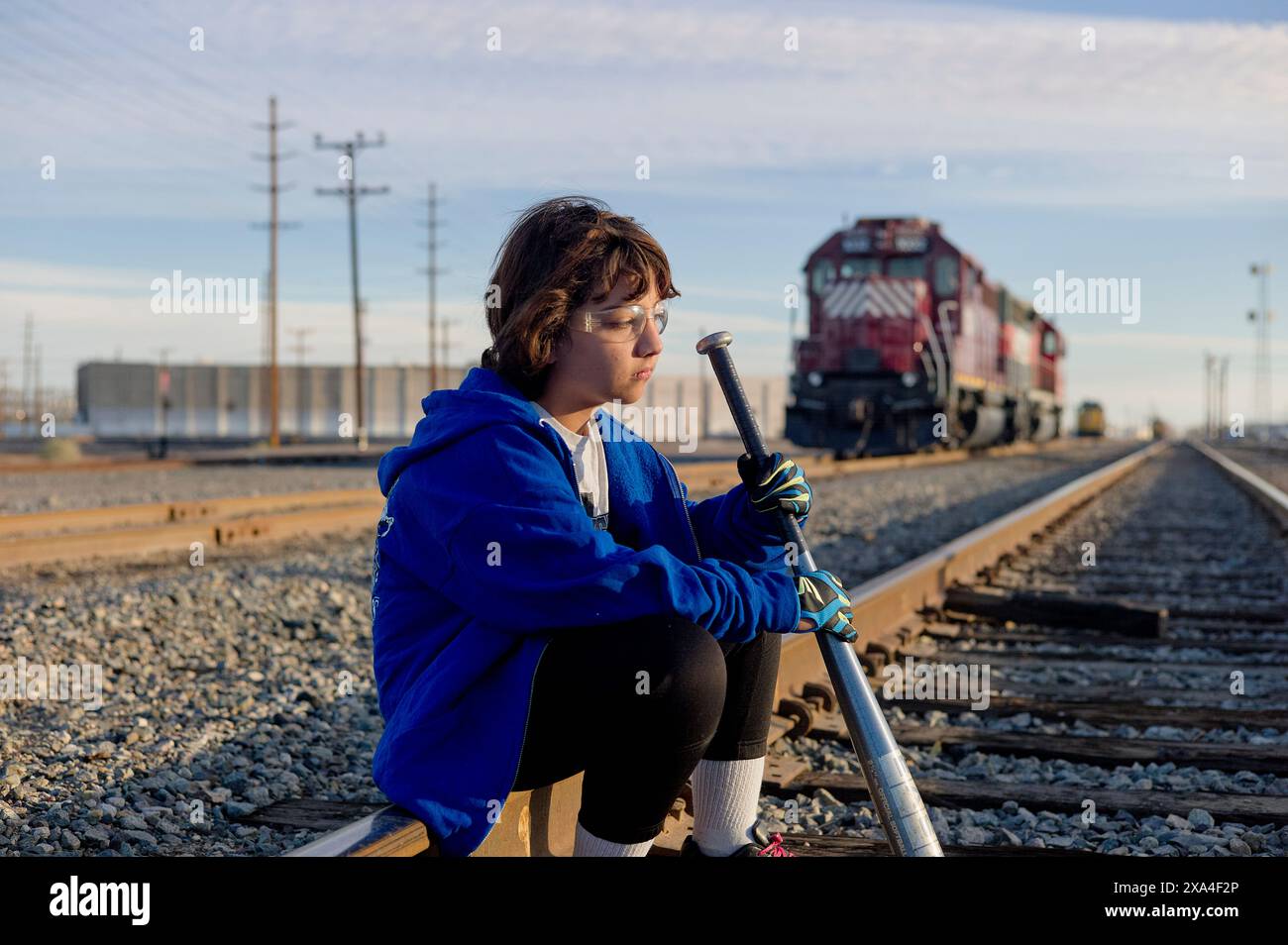 Una persona siede su un binario ferroviario, tenendo un lungo palo, con un treno e linee elettriche sullo sfondo durante il giorno. Foto Stock