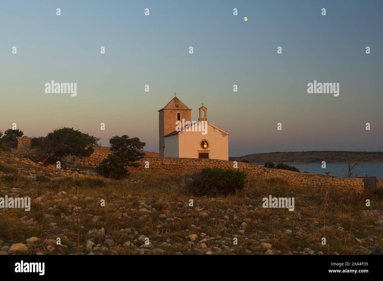 Foto dell'ultima luce del sole nella finestra della piccola chiesa cattolica sulla riva del mare Adriatico al tramonto con la luna sopra in Dalmazia, Croazia Foto Stock