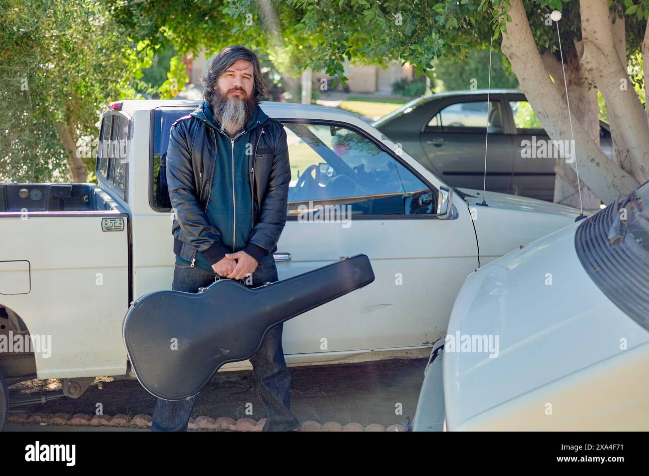 Un uomo con la barba si trova accanto a un pick-up bianco con una cassa per chitarra in una giornata di sole con alberi sullo sfondo. Foto Stock