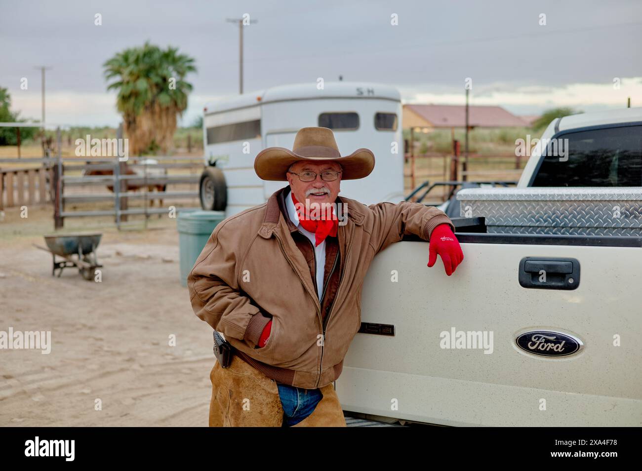Un uomo sorridente con un cappello da cowboy e guanti rossi appoggiato su un pick-up Ford bianco con un rimorchio per cavalli sullo sfondo e un ambiente rurale. Foto Stock