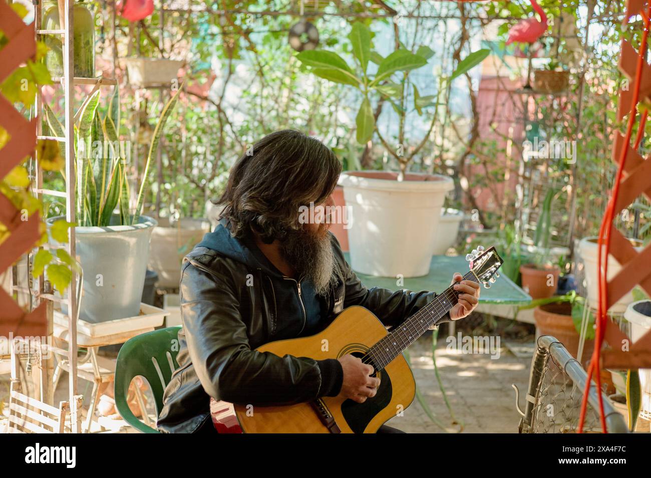 Un uomo barbuto dai capelli lunghi suona una chitarra acustica in un accogliente giardino circondato da piante in vaso e oggetti decorativi. Foto Stock