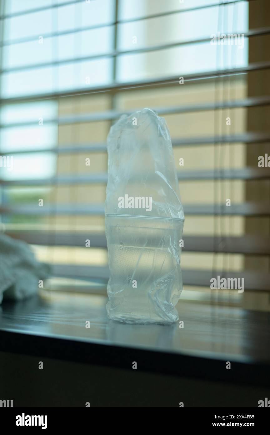 Una bottiglia di plastica traslucida e schiacciata si trova su una superficie scura sullo sfondo di una finestra con tendine orizzontali, consentendo il filtraggio della luce solare diffusa. Foto Stock