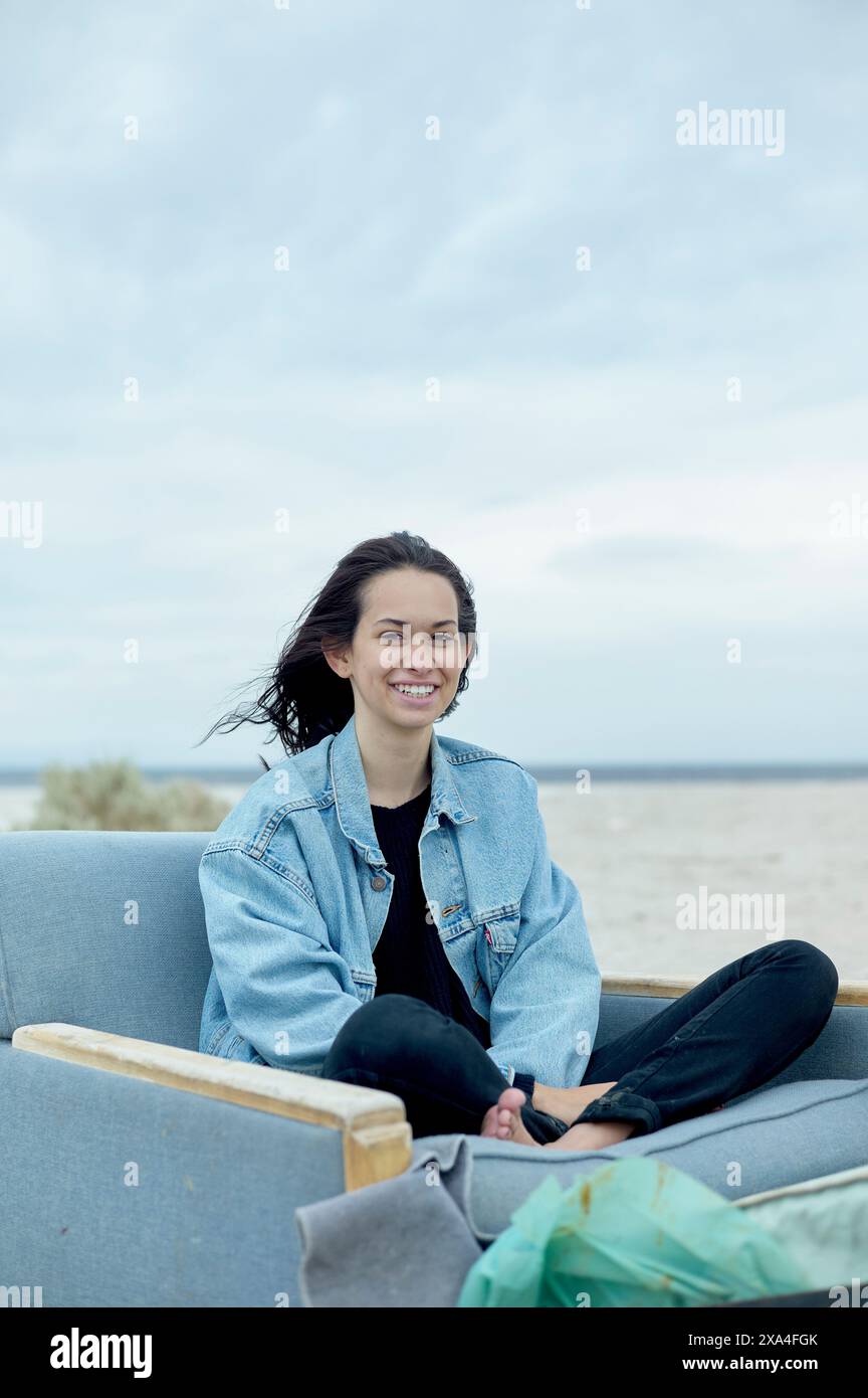 Una donna sorridente si siede comodamente su un divano esterno grigio, indossando una giacca in denim e pantaloni neri, con una spiaggia sabbiosa e un cielo nuvoloso sullo sfondo. Foto Stock