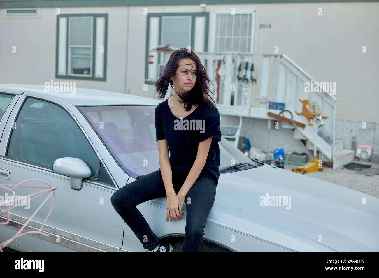 Una donna siede sul cofano di una vecchia auto d'argento di fronte a un edificio con una scala e vari oggetti disseminati intorno. Foto Stock