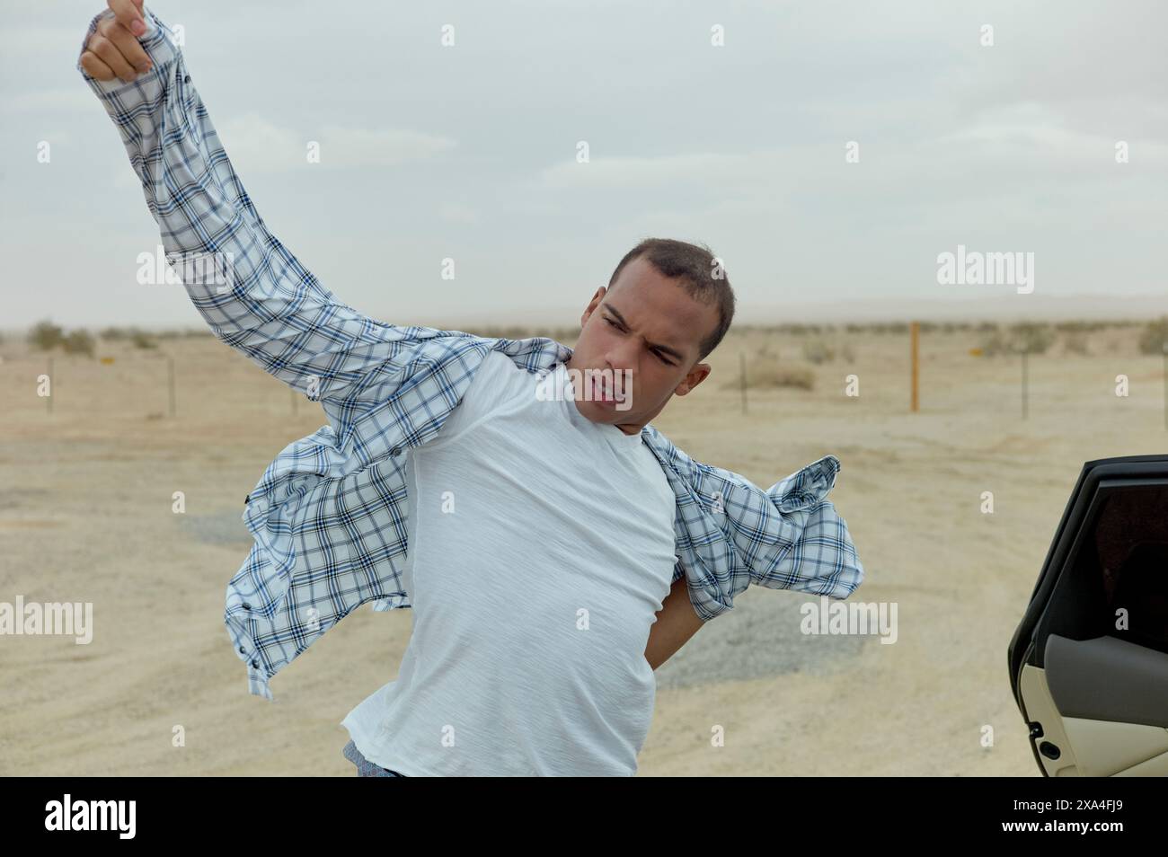 Un uomo è mezzo all'interno di un'auto parcheggiata in un'area desertica, indossando o regolando energicamente una camicia a scacchi blu e bianca. Foto Stock