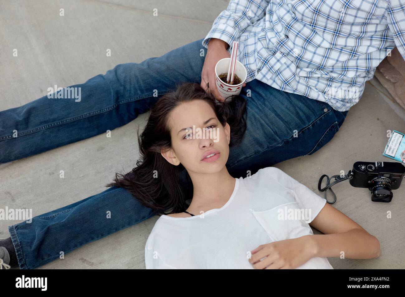 Una donna è sdraiata sulle ginocchia di un uomo per terra, sorseggiando un drink e guardando la fotocamera, con una fotocamera e uno smartphone accanto. Foto Stock