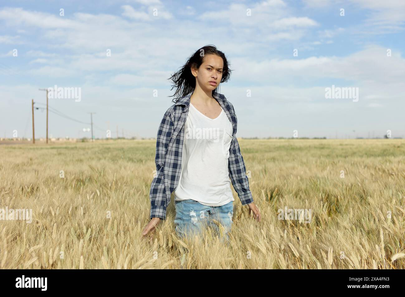 Una persona sta in mezzo a un campo di grano con un'espressione seria, indossando una canotta bianca, una camicia a scacchi e jeans blu. Foto Stock