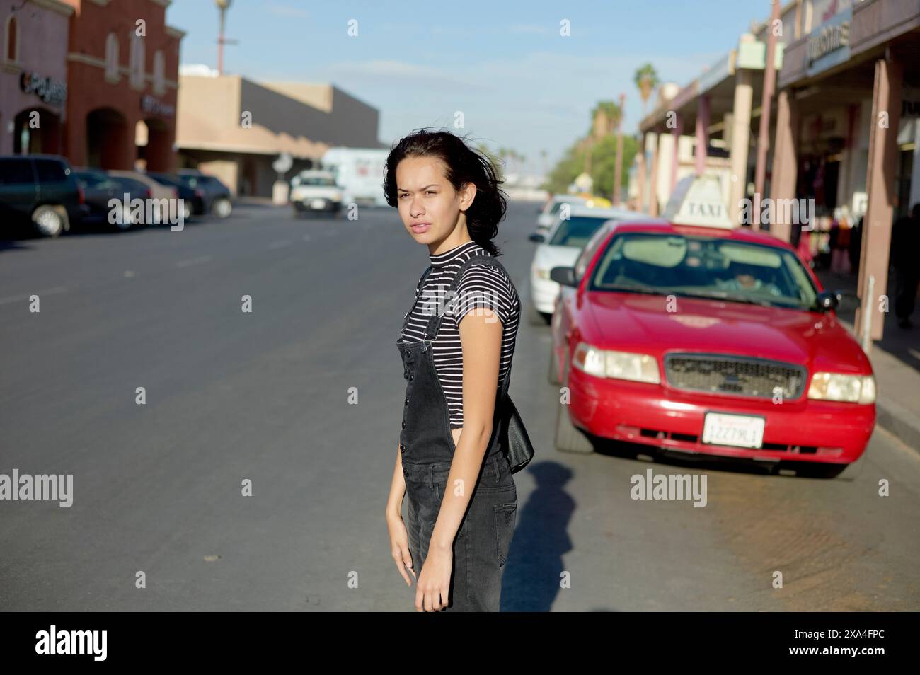 Una donna si trova in una strada della città con negozi sullo sfondo e un'auto rossa parcheggiata lateralmente. Foto Stock