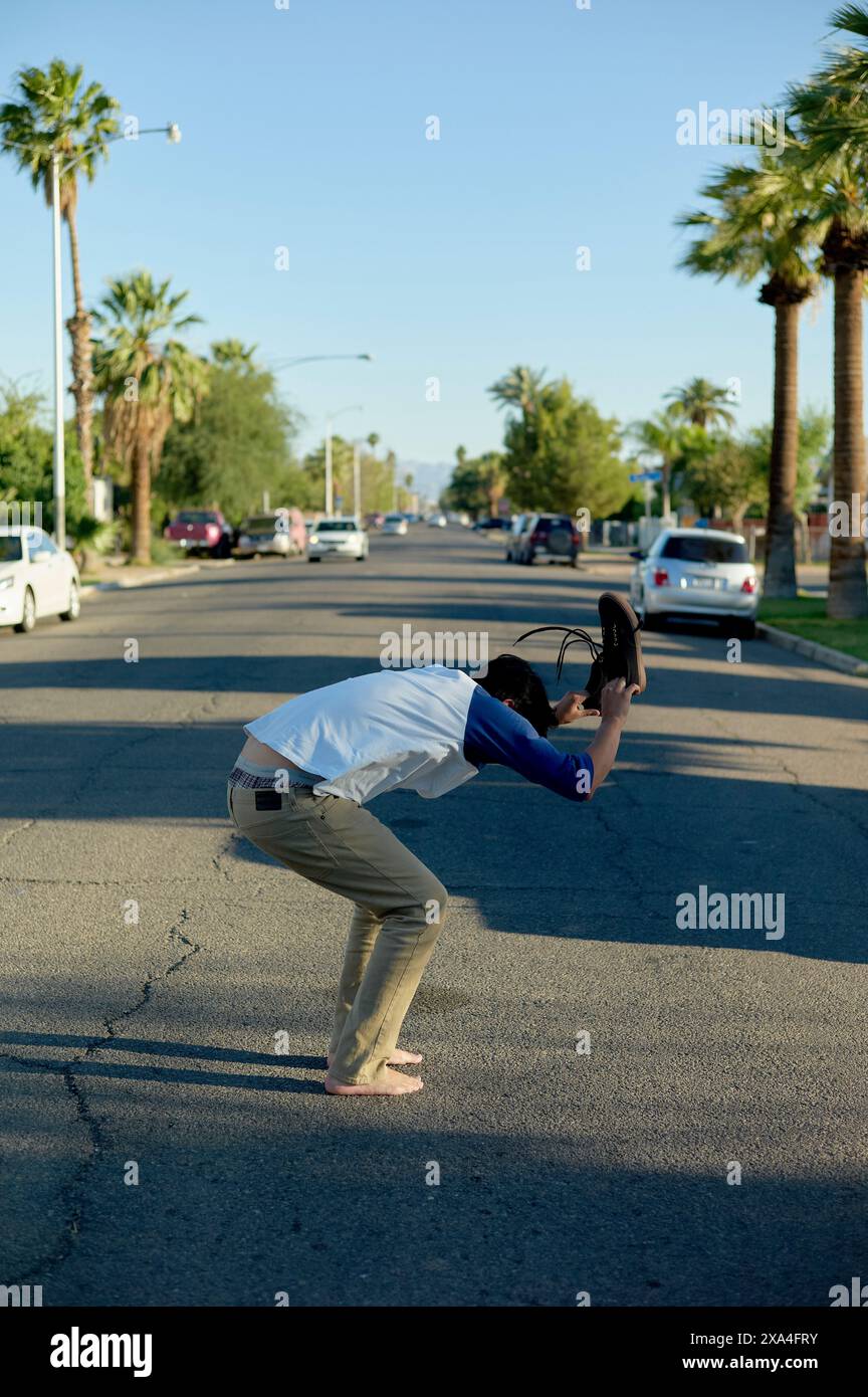 Una persona si sta piegando in avanti nel mezzo di una strada alberata, tenendo una sedia nera sopra la testa con entrambe le mani. Foto Stock