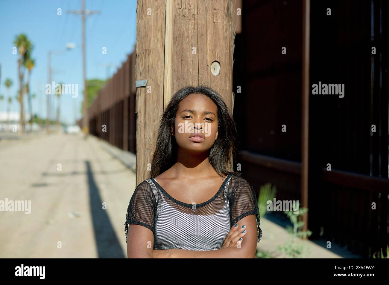 Una giovane donna si trova in un ambiente di strada, appoggiata su un palo di legno con le braccia incrociate. Foto Stock