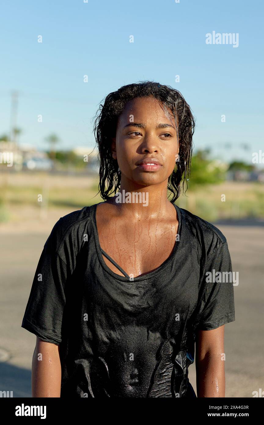 Una donna sta all'aperto con i capelli e la camicia nera bagnata, presumibilmente dall'acqua, guardando direttamente la fotocamera sotto la luce del sole. Foto Stock