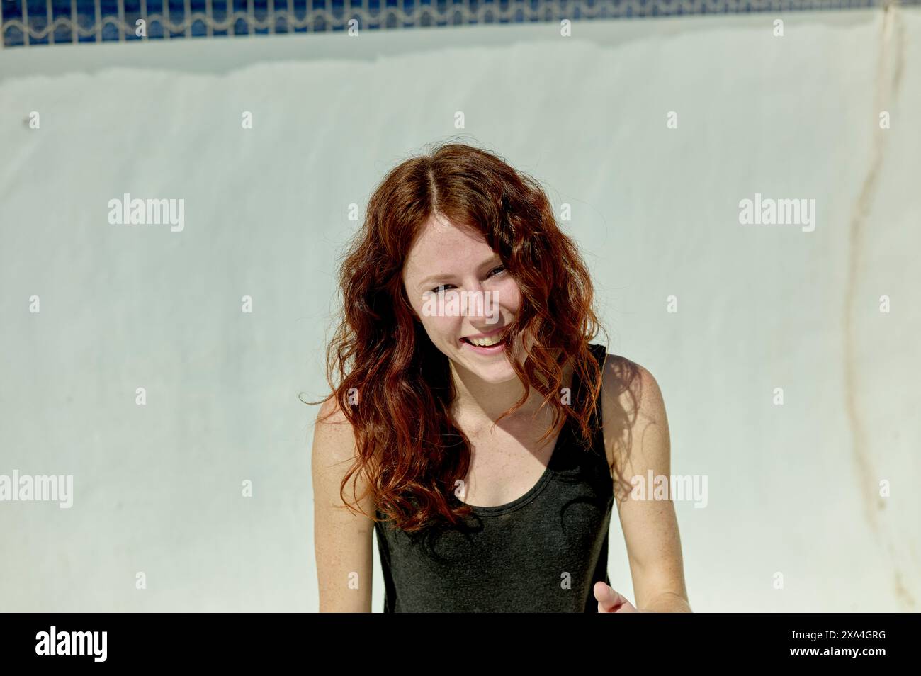 Una giovane donna allegra con lunghi capelli rossi si trova accanto a una piscina, il suo sorriso che trasmette un momento di gioia o di risate alla luce del sole. Foto Stock