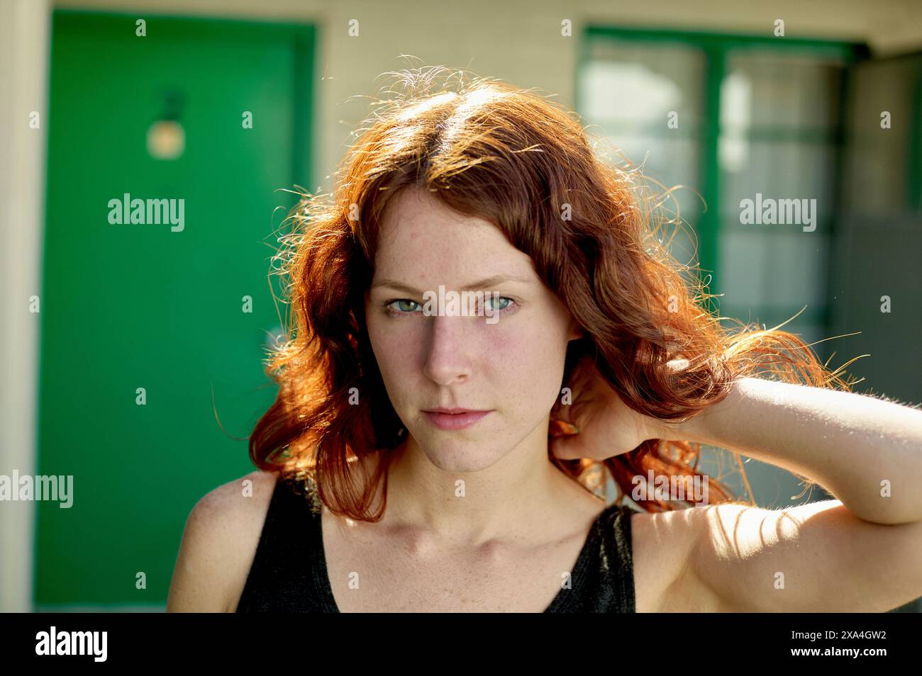 Una donna dai capelli rossi con occhi verdi sorprendenti è pronta davanti a una porta verde, la sua mano che corre tra i capelli. Foto Stock