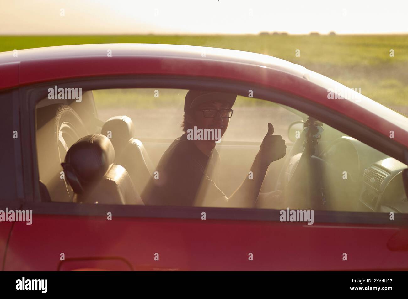 Una persona sorridente in un'auto rossa regala un pollice attraverso la finestra aperta, con un tramonto dorato sullo sfondo. Foto Stock