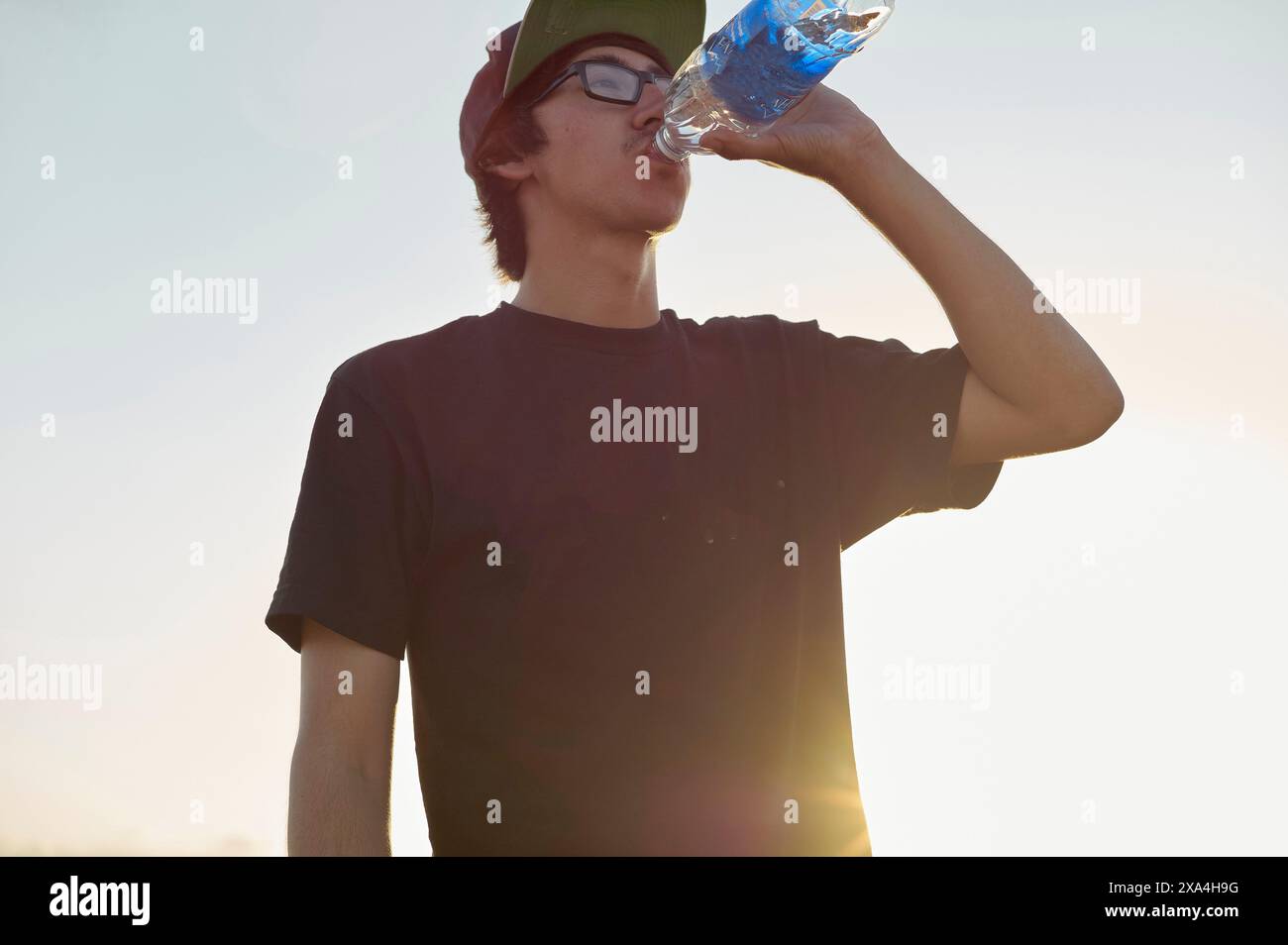 Un giovane maschio adulto beve acqua da una bottiglia blu, illuminata dalla luce del sole con un cielo limpido sullo sfondo. Foto Stock