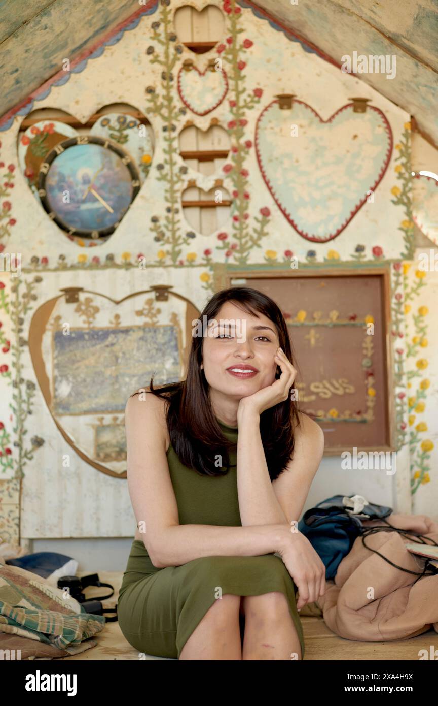 Una donna sorridente è seduta al chiuso con il mento appoggiato sulla mano, indossando un abito senza maniche verde. Lo sfondo presenta forme cardiache e motivi floreali. Foto Stock