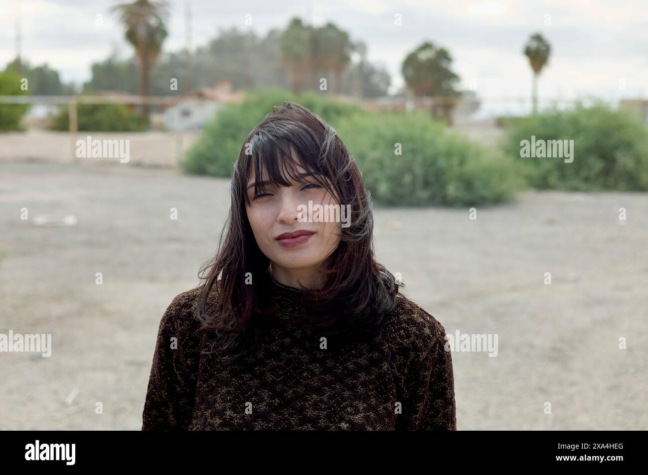 Una donna si staglia all'aperto con i capelli spazzati dal vento, indossando un maglione a motivi scuri e un'espressione delicata su uno sfondo sfocato di vegetazione ed edifici. Foto Stock