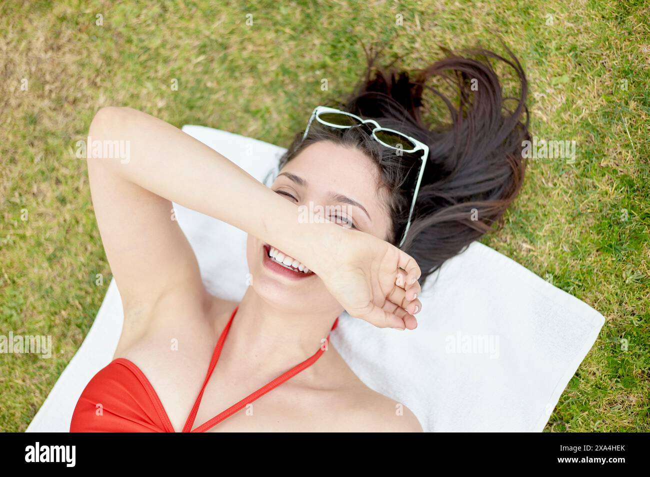 Una donna gioiosa giace sull'erba verde, indossa un costume da bagno rosso con occhiali da sole bianchi sulla testa, coprendo gli occhi con la mano. Foto Stock