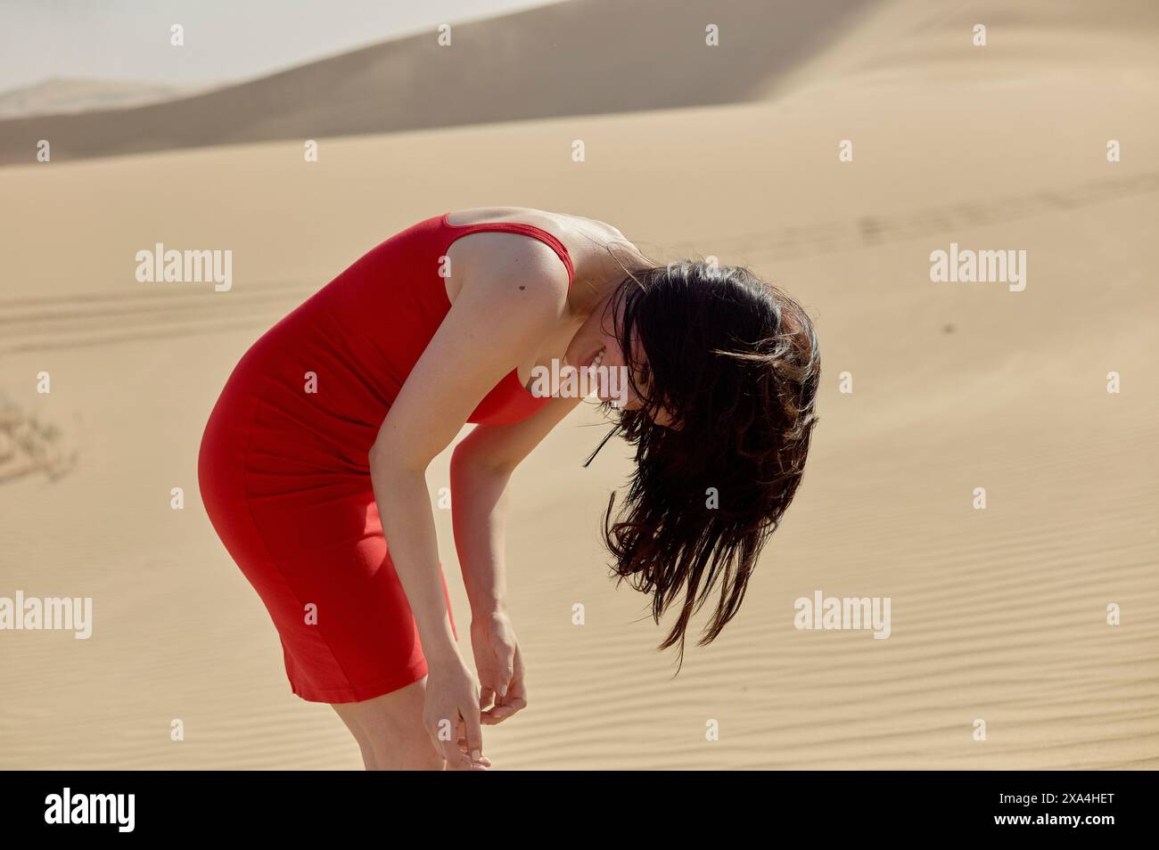 Una donna con un vestito rosso si piega in avanti e sorride, in piedi tra le dune di sabbia. Foto Stock
