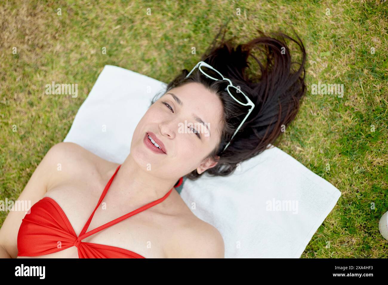 Una donna è sdraiata sulla schiena su un asciugamano bianco posto sull'erba, indossa un costume da bagno rosso e occhiali da sole sulla testa, con un sorriso rilassato sul viso. Foto Stock