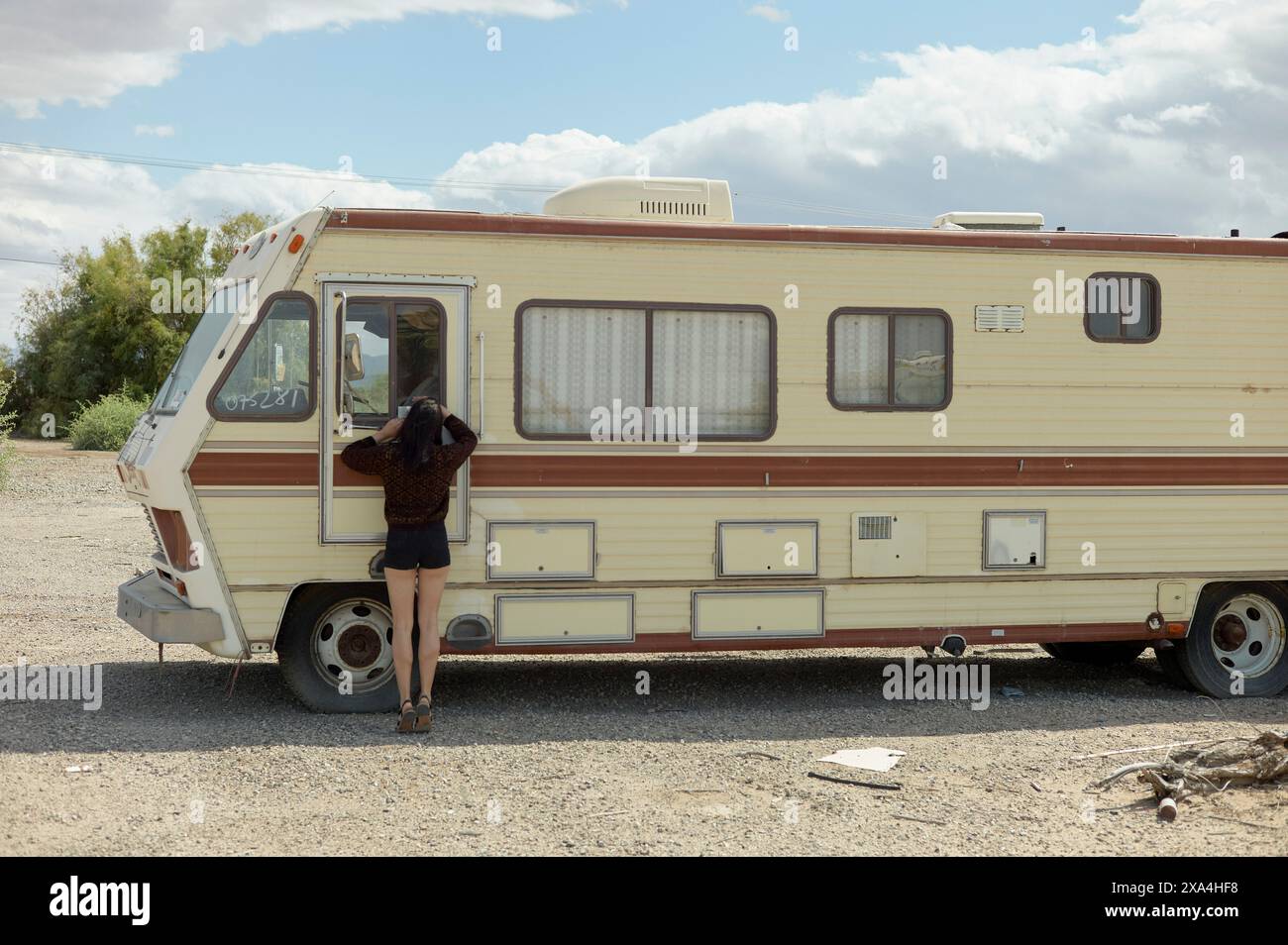Un individuo sta sbirciando in un camper d'epoca parcheggiato su un tranquillo terreno desertico sotto un cielo parzialmente nuvoloso. Foto Stock