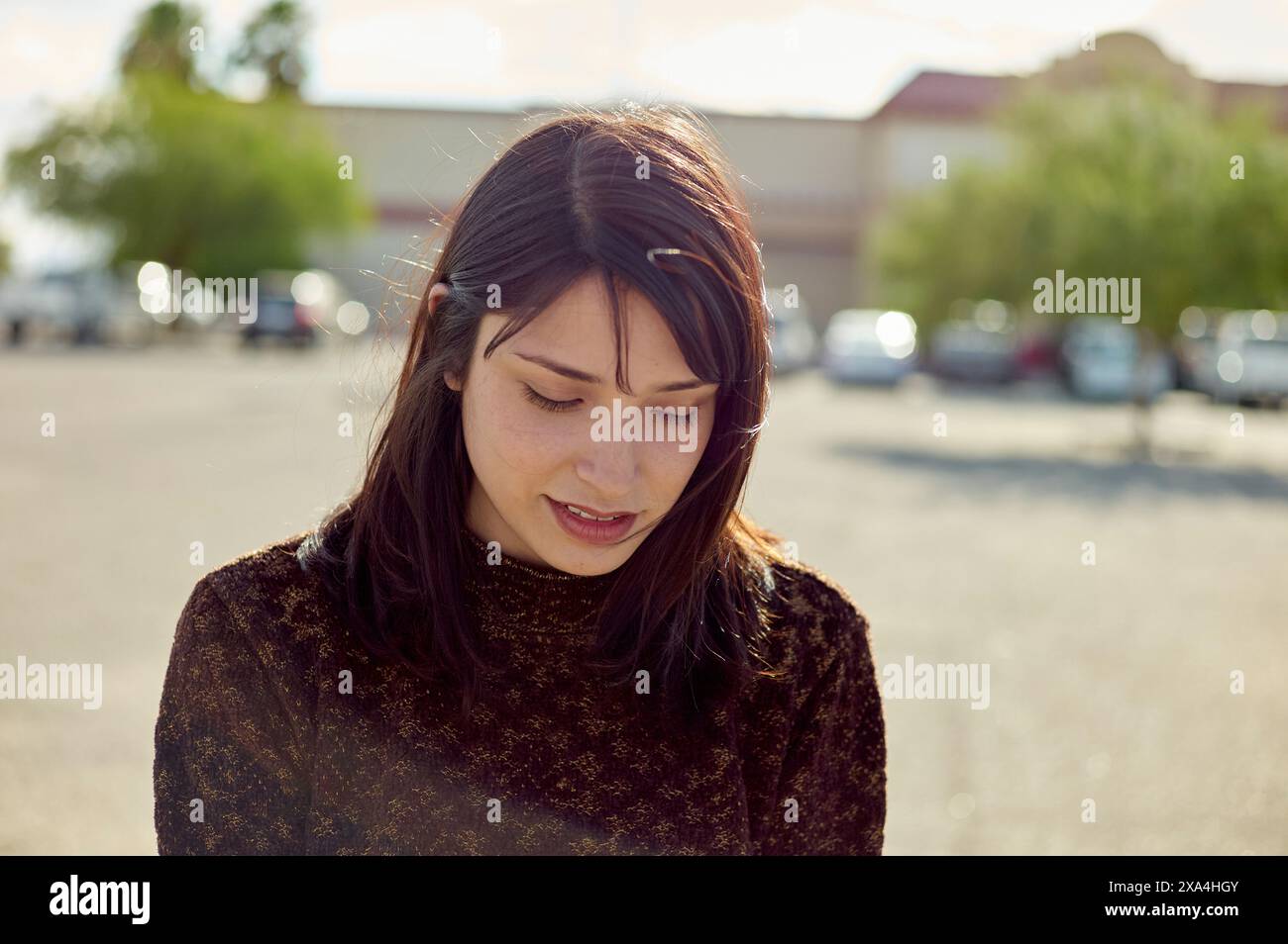 Una donna con i capelli scuri sta all'aperto in una giornata di sole, il suo sguardo è rivolto verso il basso mentre la luce del sole filtra i capelli. Foto Stock