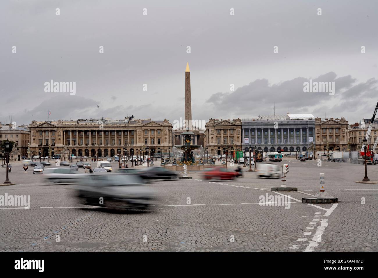 L'immagine raffigura una vivace piazza urbana con un obelisco e un edificio classico sullo sfondo sotto un cielo coperto. I veicoli sono in movimento, sfocati in primo piano. Foto Stock