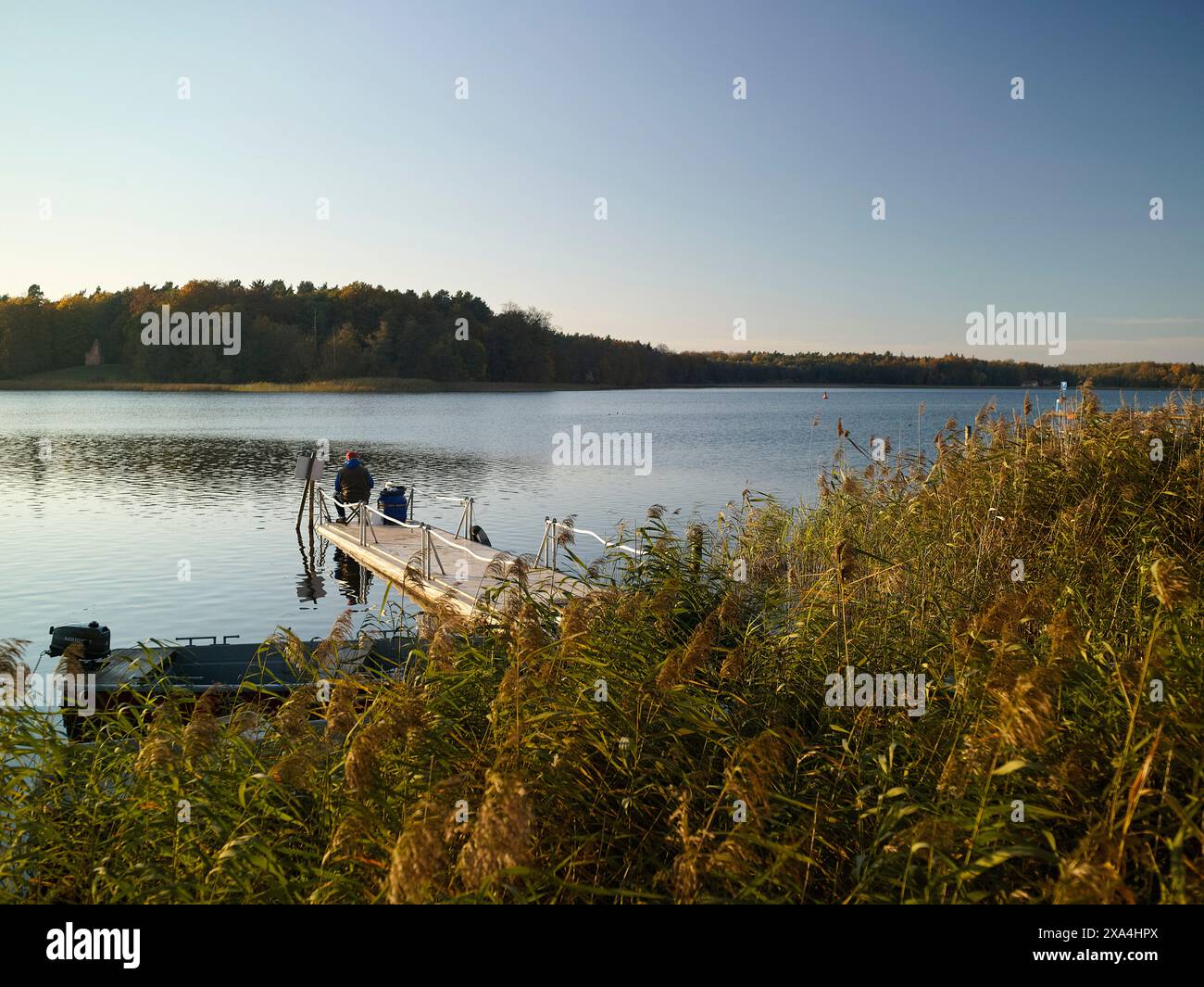 Un tranquillo lago con due persone sedute su un molo durante il giorno, circondato da acque calme e lussureggiante vegetazione sotto un cielo limpido. Foto Stock