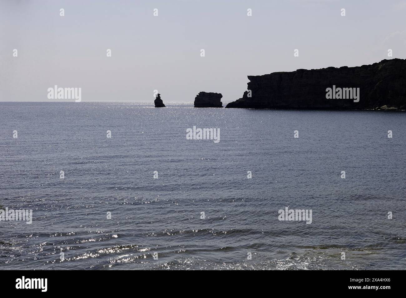 Paesaggio costiero caratterizzato dal mare con dolci onde in primo piano e una scogliera rocciosa sulla destra. Una formazione rocciosa indipendente è visibile in lontananza vicino al bordo della scogliera. Foto Stock