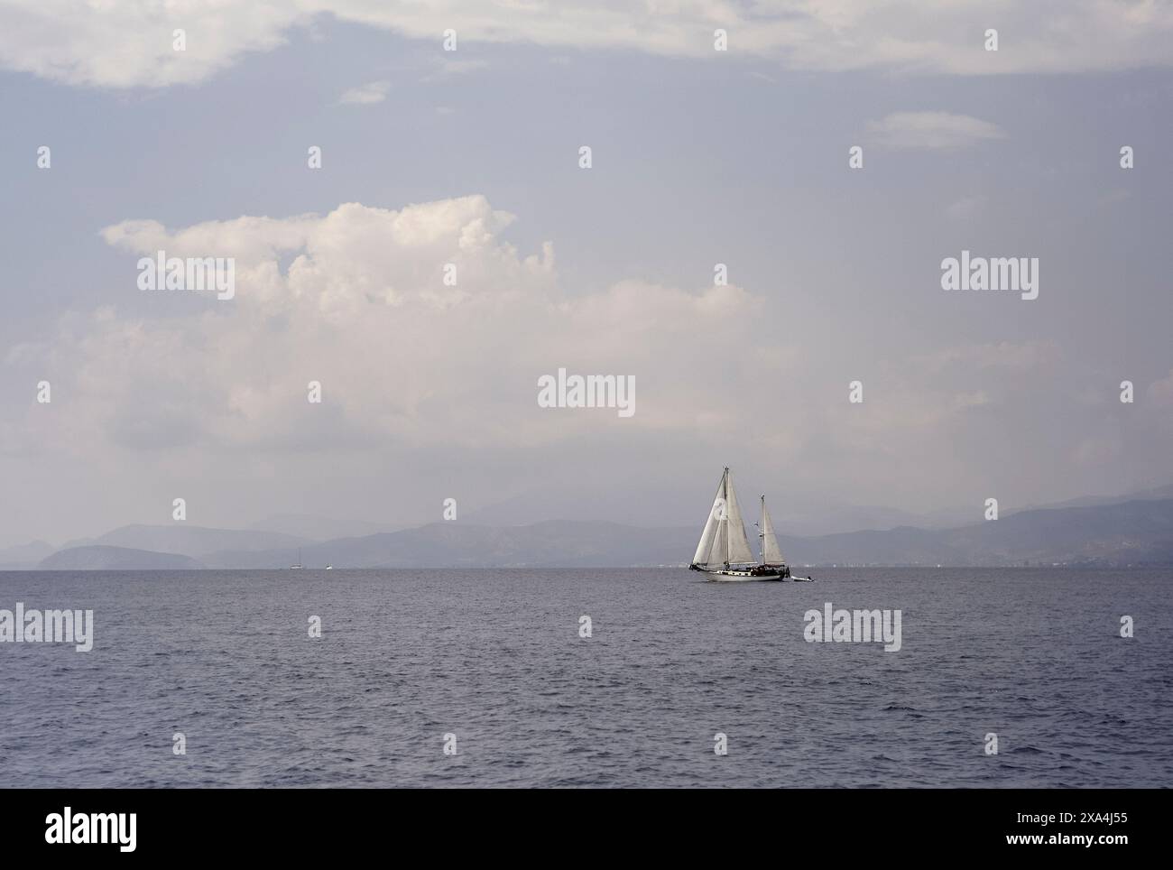 Una barca a vela scivola sulle acque calme sotto un cielo nuvoloso con montagne lontane all'orizzonte. Foto Stock
