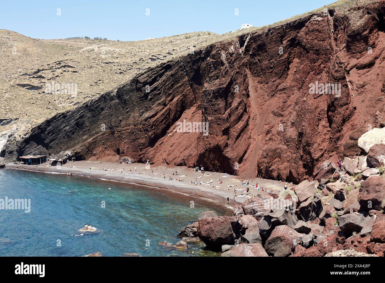 Una tranquilla spiaggia di sabbia rossa con gente che prende il sole e nuota, circondata da scogliere scoscese e scoscese sotto un cielo azzurro. Foto Stock