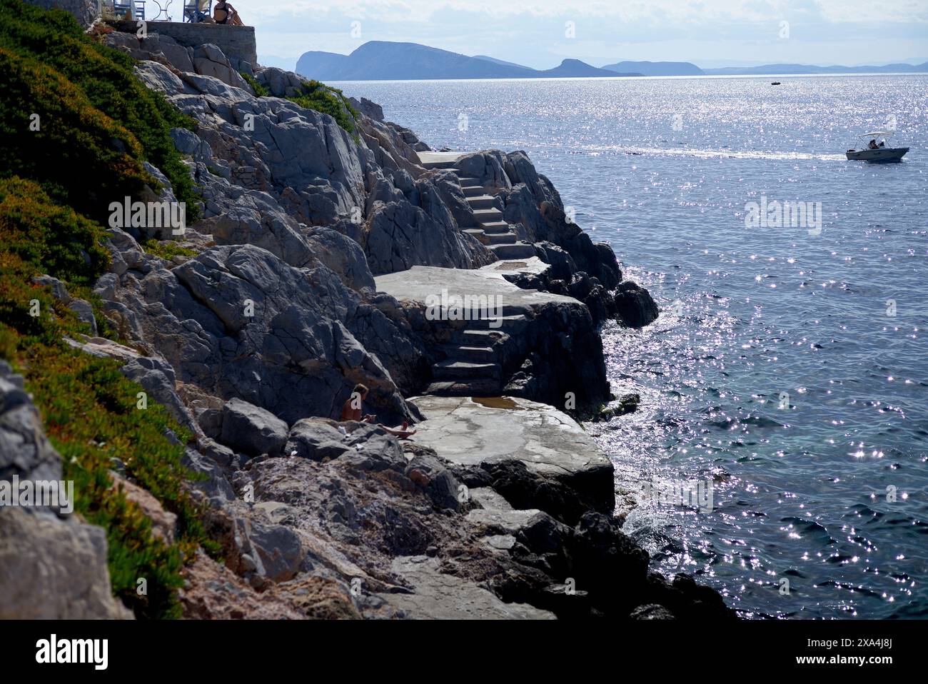 Un'aspra scalinata in pietra scende lungo una scogliera verso il mare, con persone che si godono la vista e una barca in lontananza. Foto Stock