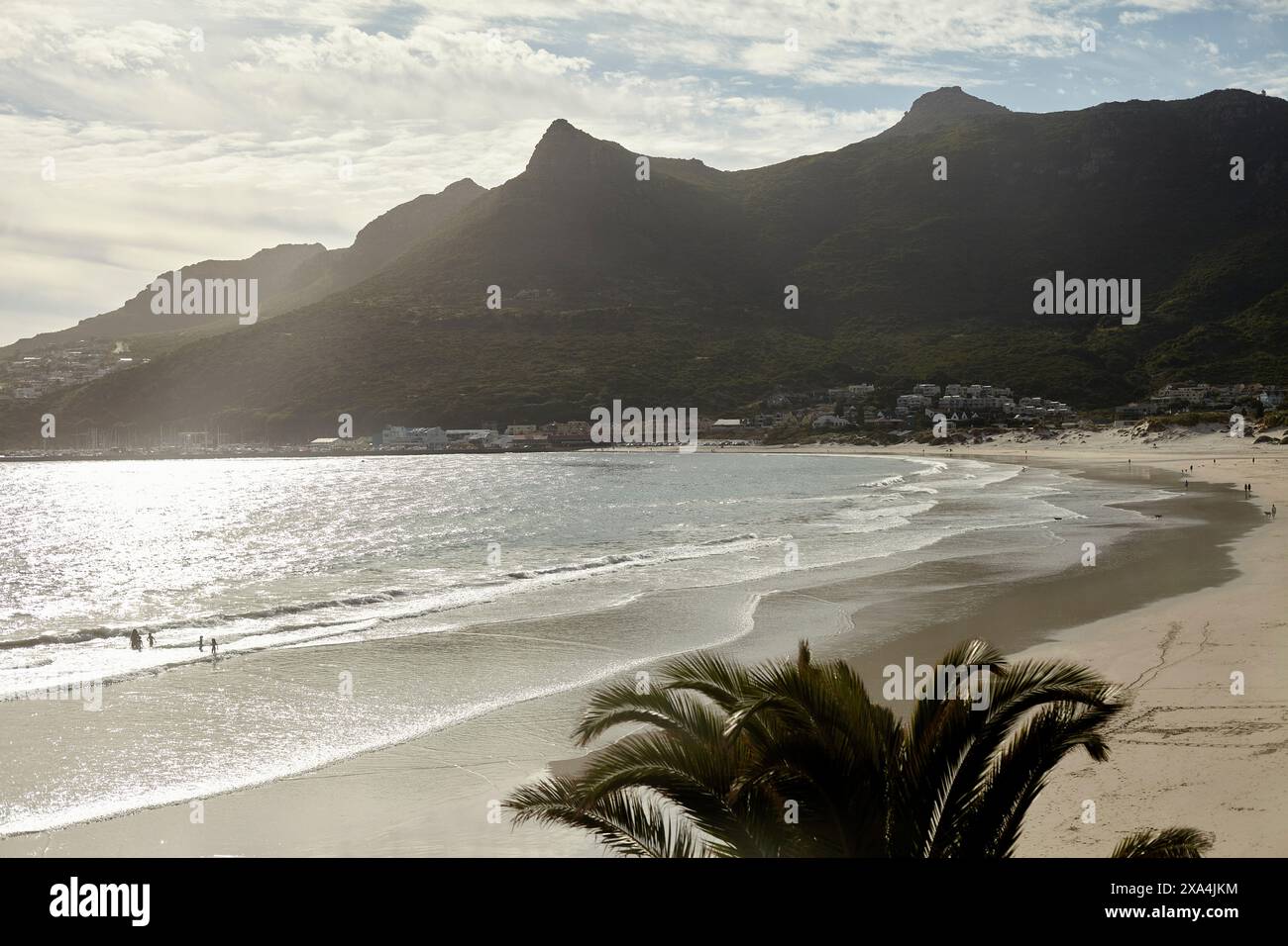 Una vista mozzafiato di una spiaggia tranquilla con uno sfondo di montagna e un cielo limpido. Il sole splende dall'acqua mentre la gente si gode il mare tranquillo. Foto Stock