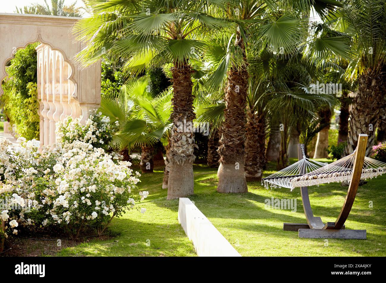 Un tranquillo giardino con vegetazione lussureggiante, un'amaca adagiata tra palme, fiori bianchi in fiore e un muro con caratteristiche architettoniche tradizionali. Foto Stock