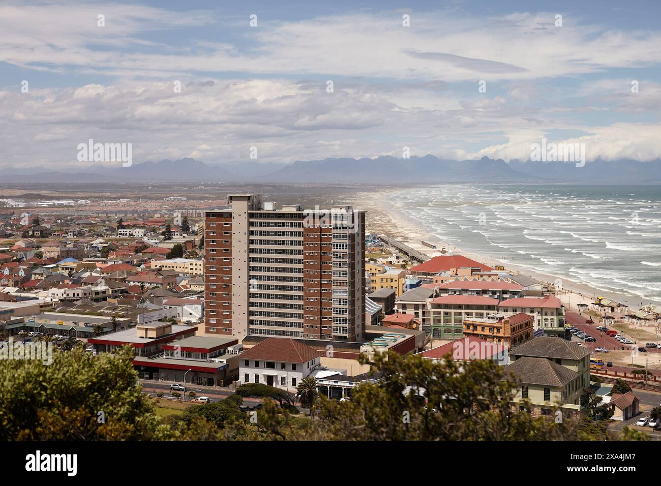 Una vista panoramica di una città costiera con un alto edificio che si erge prominentemente tra le strutture inferiori, adagiato su uno sfondo di montagne e un cielo nuvoloso. Foto Stock