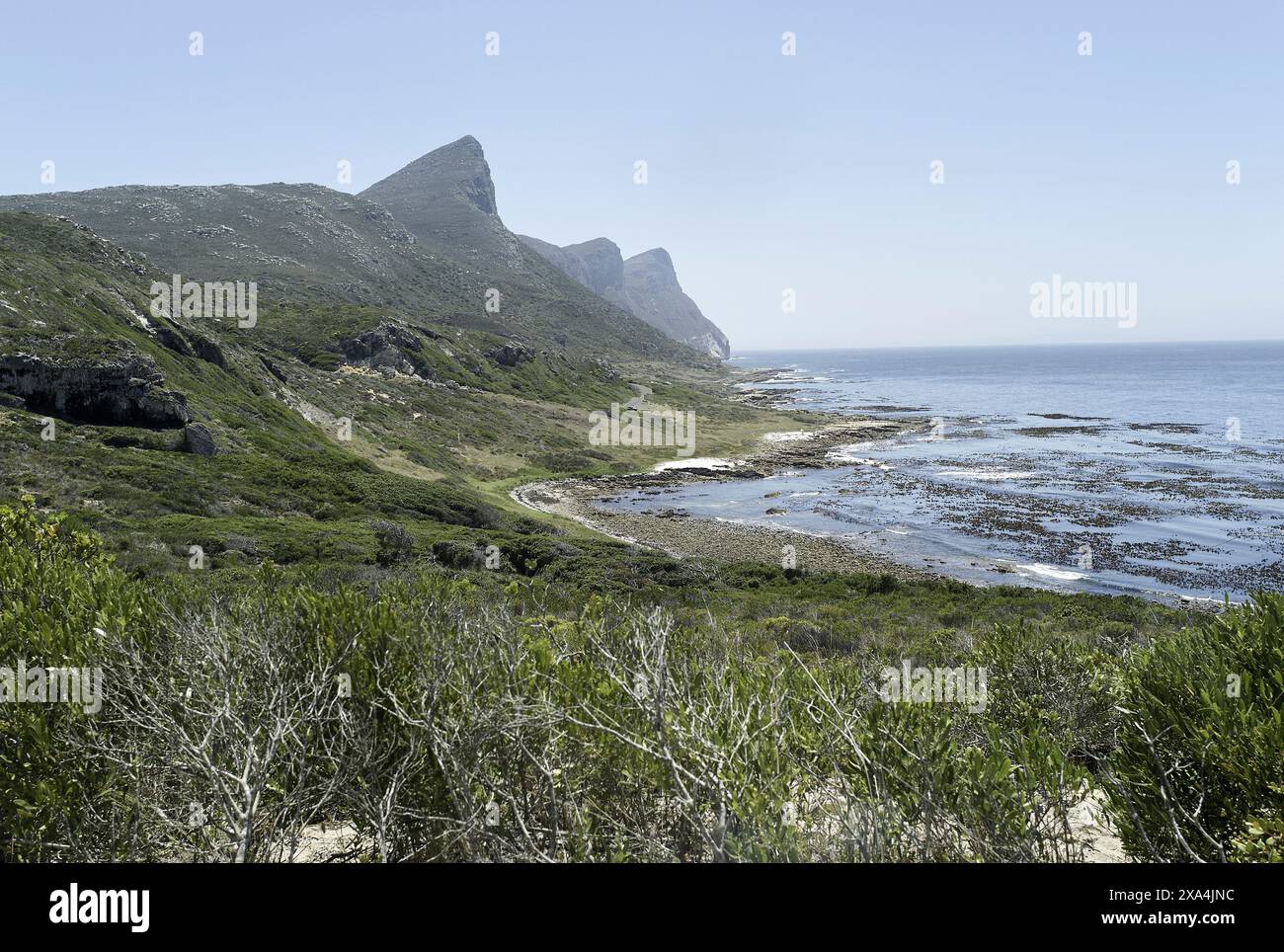 L'immagine raffigura un tranquillo paesaggio costiero con una catena montuosa sullo sfondo, una costa rocciosa e un cielo limpido. Foto Stock