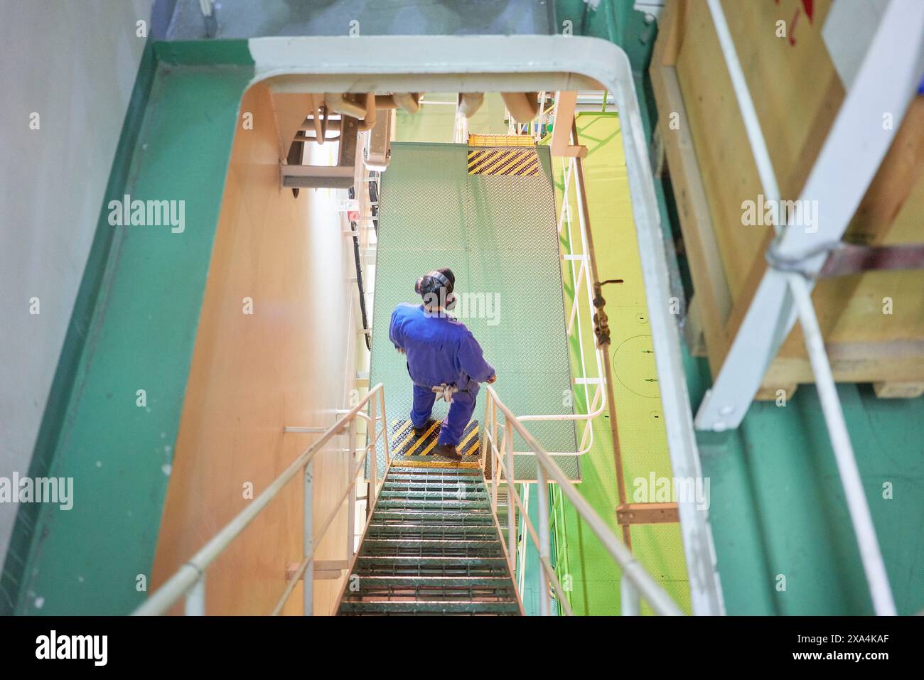 Una persona con tute blu sta scendendo una scala in metallo in un ambiente industriale con corrimano di sicurezza gialle. Foto Stock