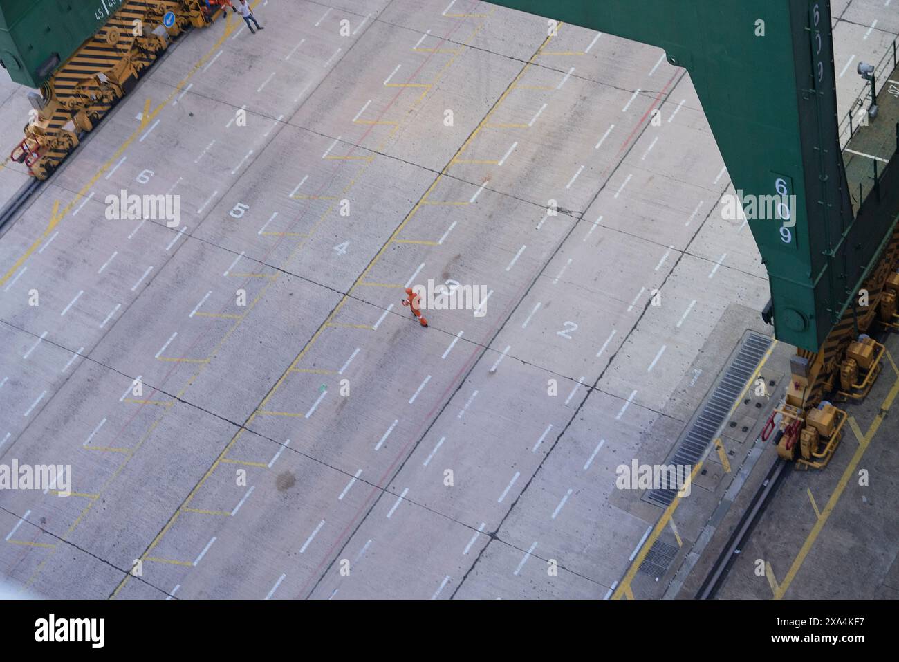 Una persona in un gilet arancione cammina attraverso un'area asfaltata segnata con linee e numeri gialli, mentre le attrezzature industriali si innalzano sulla scena e diversi individui sono visti nelle vicinanze. Foto Stock