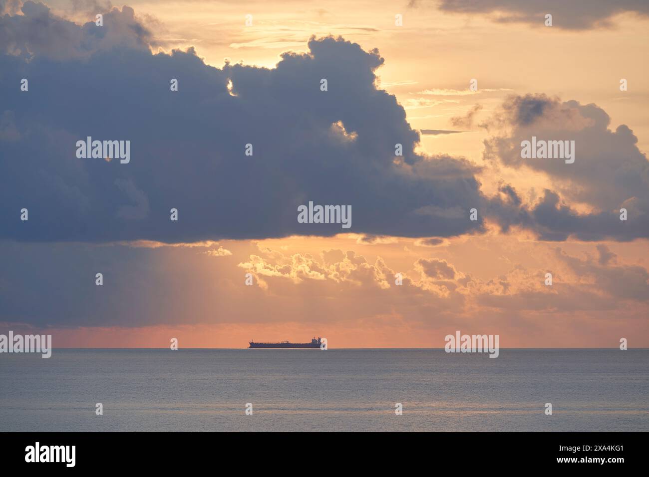 Una tranquilla scena oceanica al tramonto con una nave da carico visibile all'orizzonte, retroilluminata dalle tenui e calde sfumature del sole che sbircia attraverso nuvole sparse. Foto Stock