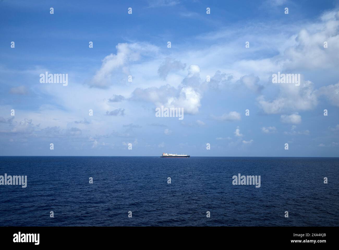 Una nave da carico siede in lontananza su un oceano blu calmo sotto un vasto cielo disseminato di nuvole bianche. Foto Stock