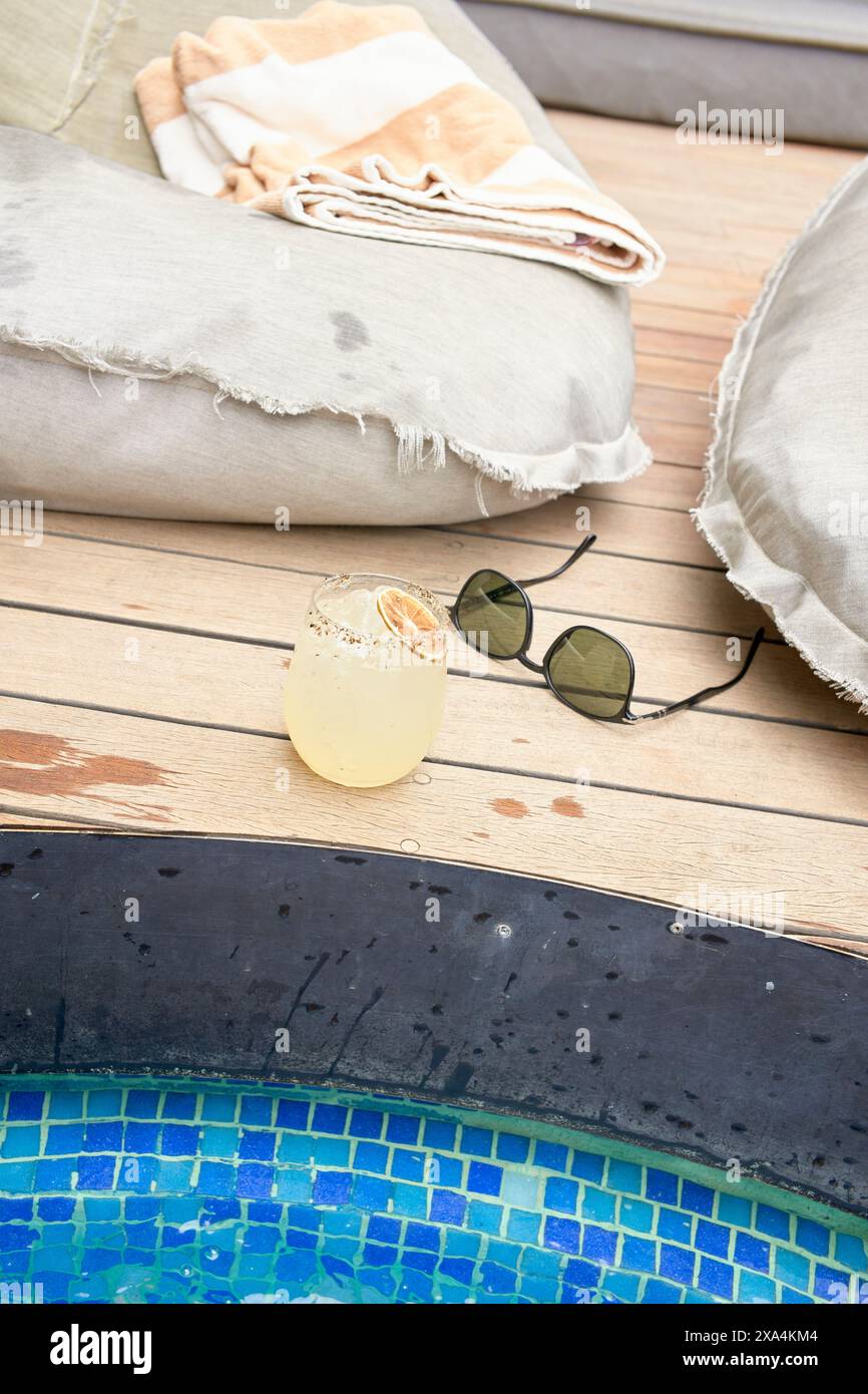 Una bevanda rinfrescante con una fetta di lime siede su una superficie di legno accanto ad una piscina, accompagnata da un paio di occhiali da sole e comodi cuscini. Foto Stock