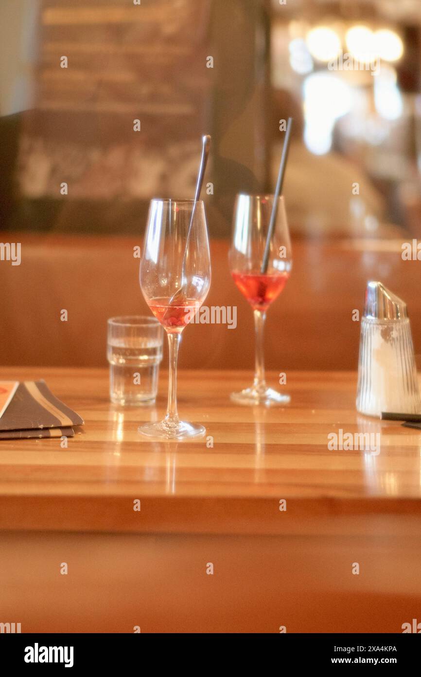 Due bicchieri da vino, uno parzialmente riempito di vino rosa, seduti su un bancone di legno del bar, accompagnati da un bicchiere d'acqua e uno shaker di sale, con uno sfondo sfocato che suggerisce un ambiente accogliente del ristorante. Foto Stock Due bicchieri da vino, uno parzialmente riempito di vino rosa, seduti su un bancone di legno del bar, accompagnati da un bicchiere d'acqua e uno shaker di sale, con uno sfondo sfocato che suggerisce un ambiente accogliente del ristorante. Foto Stock